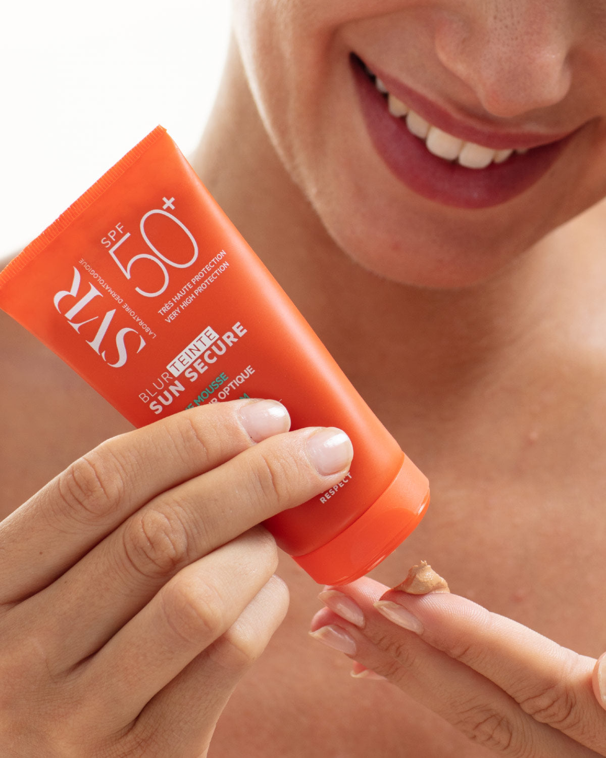 Tube orange SVR Sun Secure Blur teinté SPF50+ délivrant une texture mousse teintée sur le doigt d'une femme souriante.