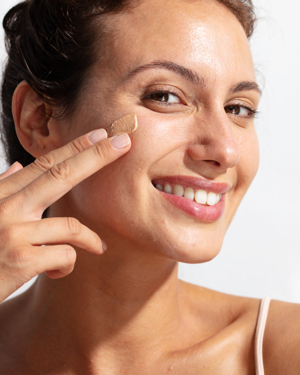 Femme souriante appliquant une crème solaire teintée beige sur sa joue pour un teint unifié et une protection SPF50+.