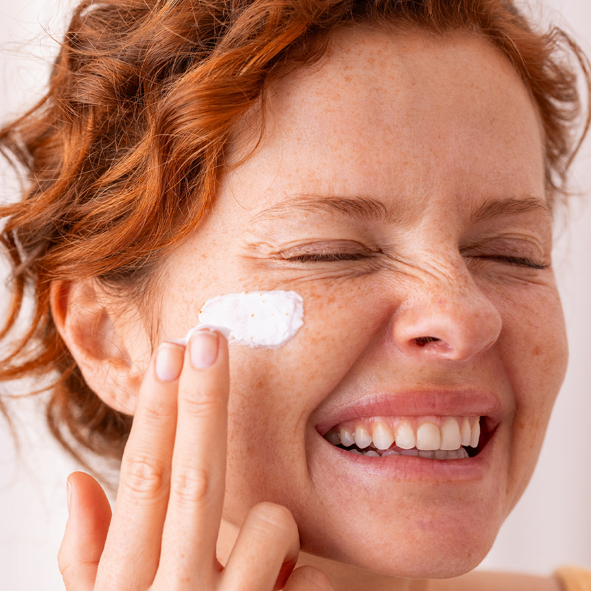 Femme rousse souriante appliquant une crème [C20] Biotic onctueuse avec microbilles vitaminées sur sa joue.