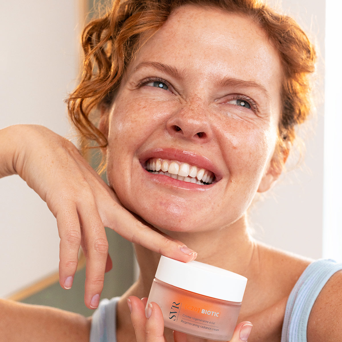Femme souriante tenant un pot de crème SVR [C20] Biotic à la texture orangée dans un emballage en verre dépoli.