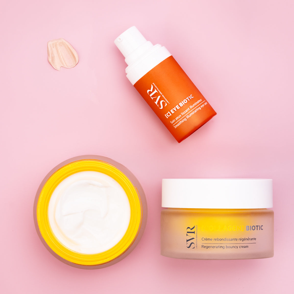 Flacon pompe orange SVR [C] EYE BIOTIC et pot de crème régénérante sur fond rose avec une touche de texture crème.