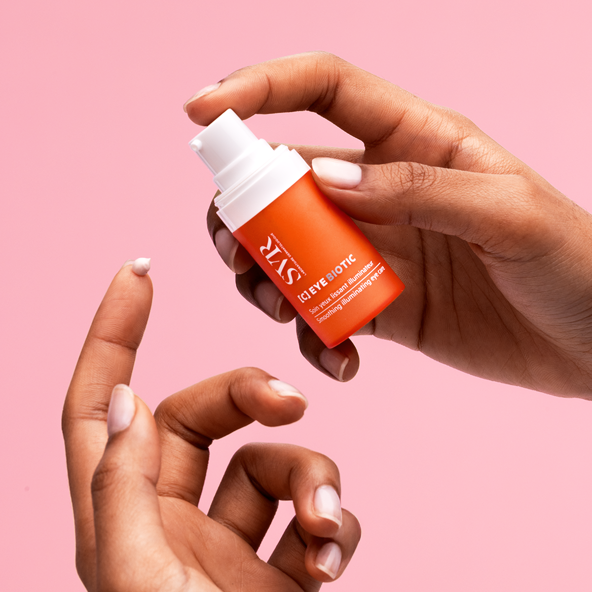 Flacon pompe orange SVR [C] EYE BIOTIC avec une noisette de crème lissante sur le doigt, sur fond rose.