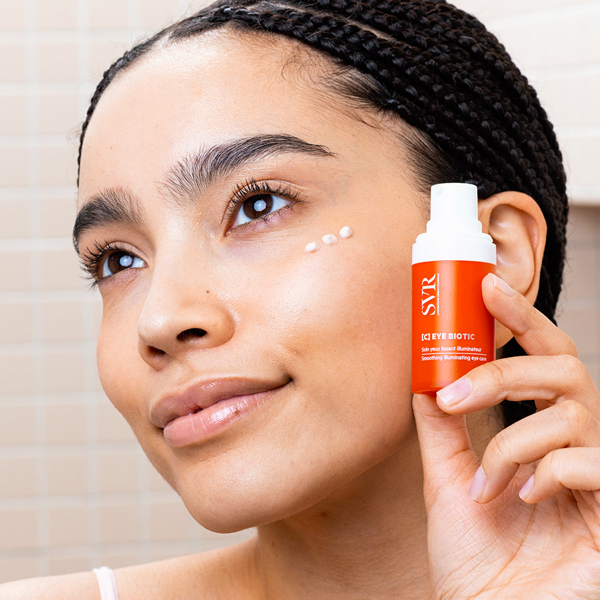 Femme tenant un flacon orange SVR [C] EYE BIOTIC avec des points de crème illuminatrice sous l'œil.