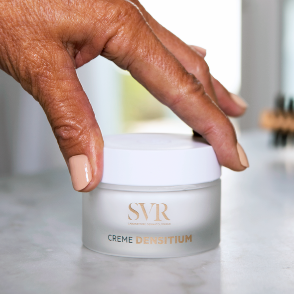 Pot de crème anti-âge SVR DENSITIUM en verre dépoli avec couvercle blanc, tenu par une main sur un support en marbre.
