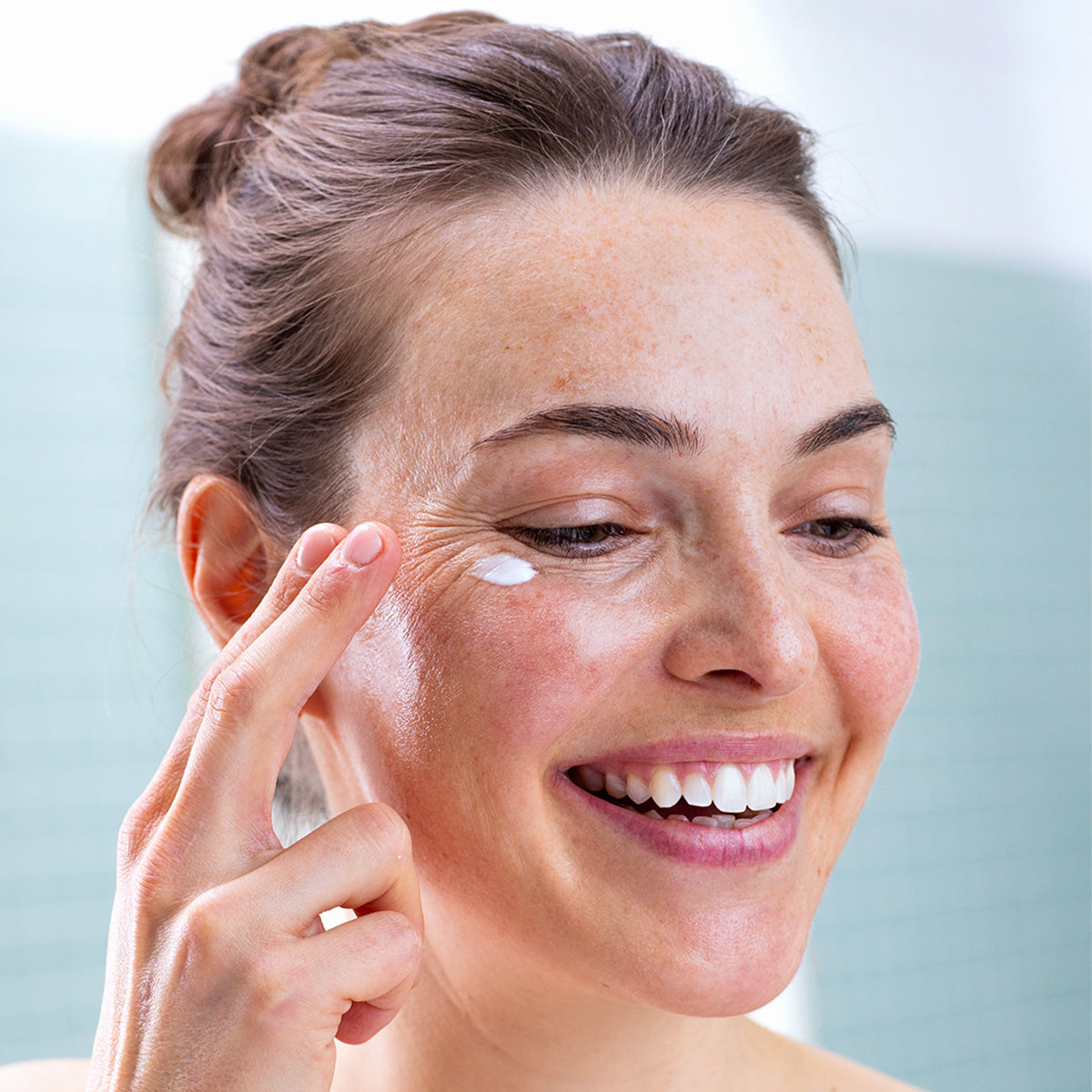 Femme souriante appliquant une noisette de crème blanche onctueuse sur le contour de ses yeux sensibles.