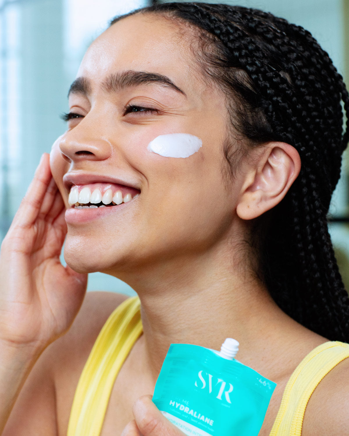 Femme souriante appliquant la crème hydratante SVR Hydraliane sur sa joue avec le nouveau format poche éco-responsable bleu.