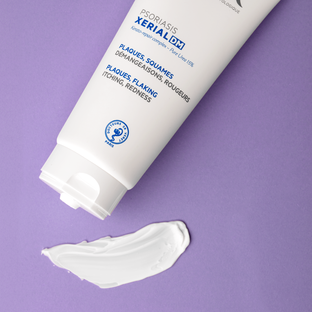 Tube blanc de crème Xerial DM pour le psoriasis avec une noisette de texture onctueuse sur fond violet.