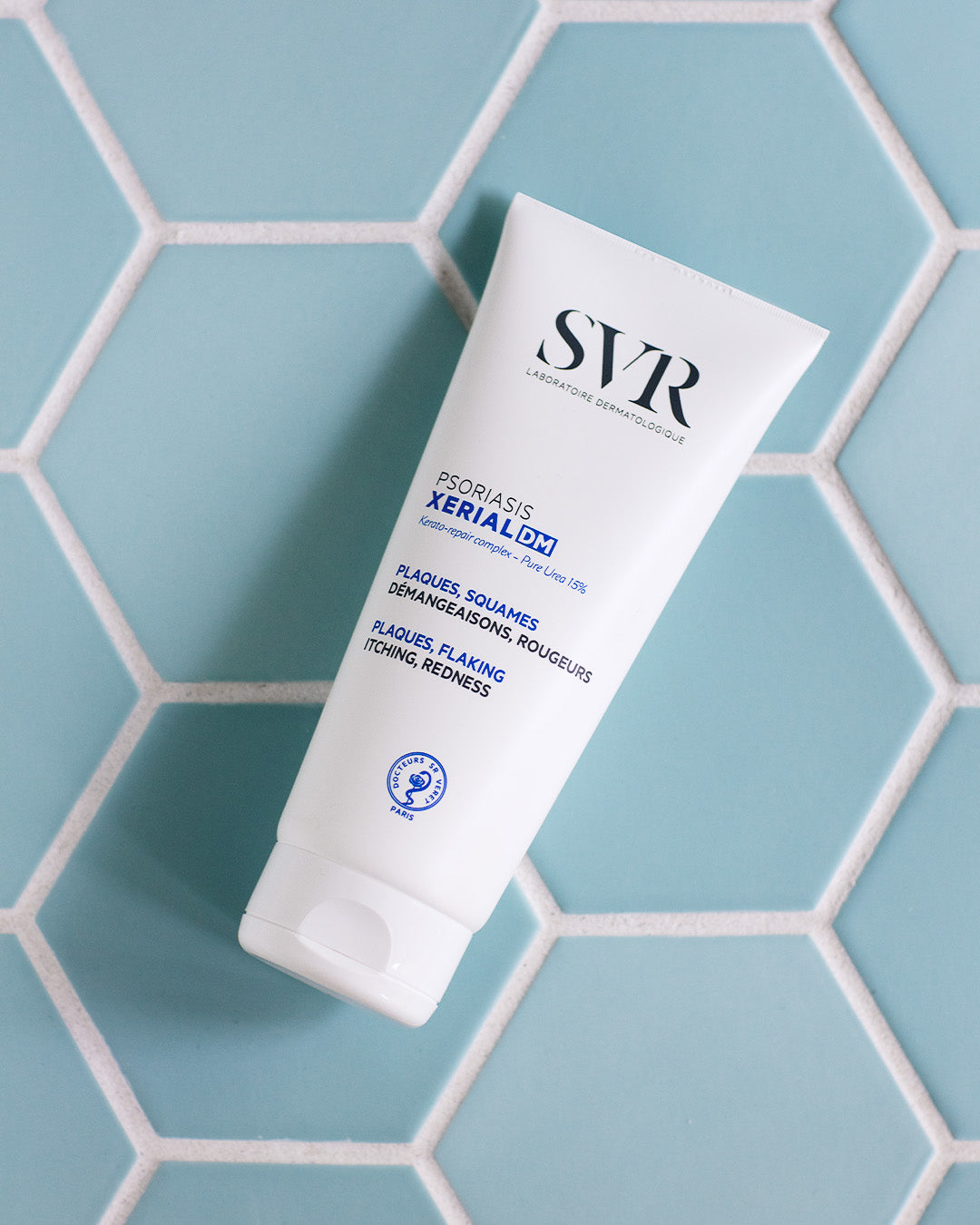 Tube de crème SVR XERIAL DM pour le psoriasis sur un fond de carrelage hexagonal bleu clair.