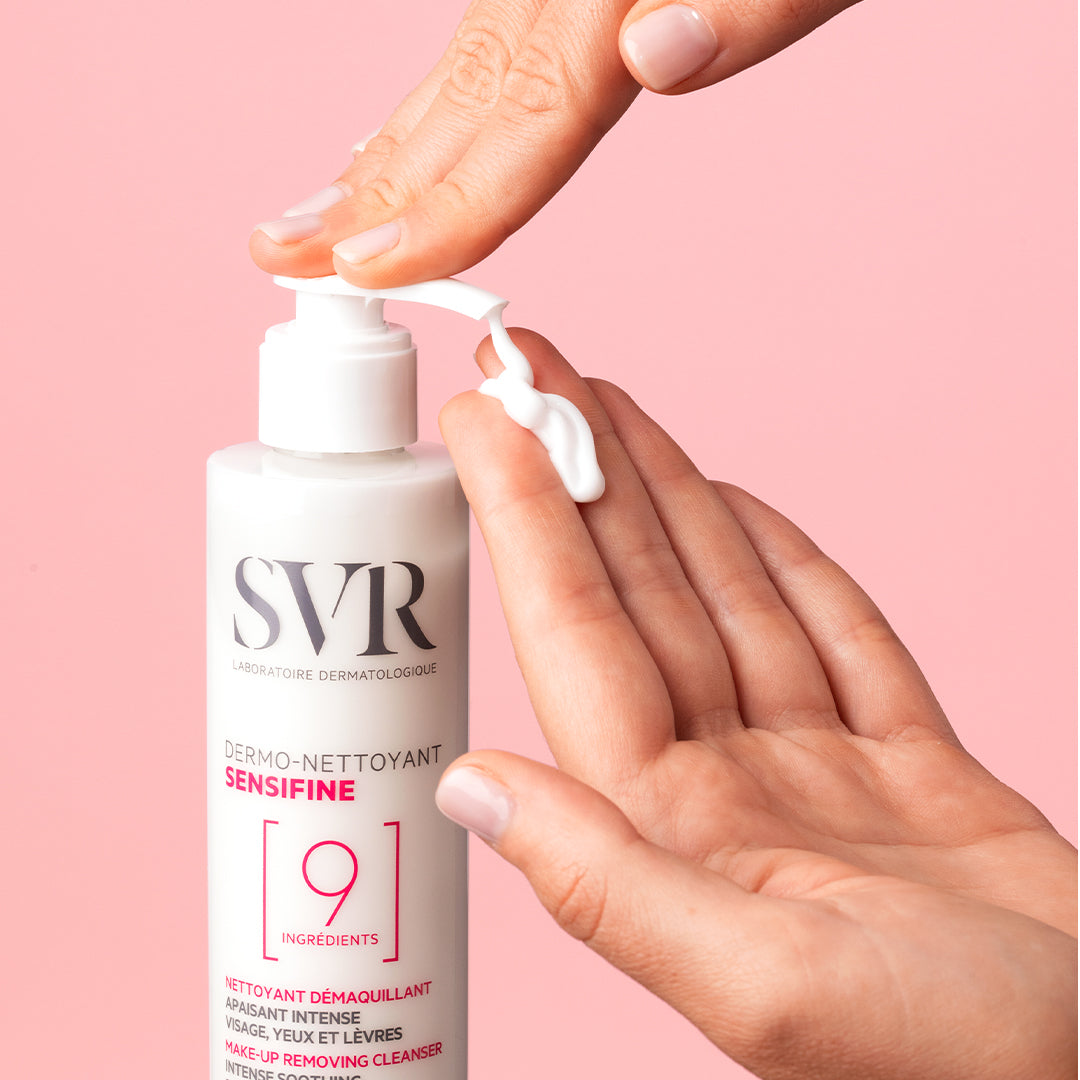 Flacon pompe SVR Sensifine Dermo-Nettoyant délivrant une texture gel-crème blanche sur une main, fond rose pastel.