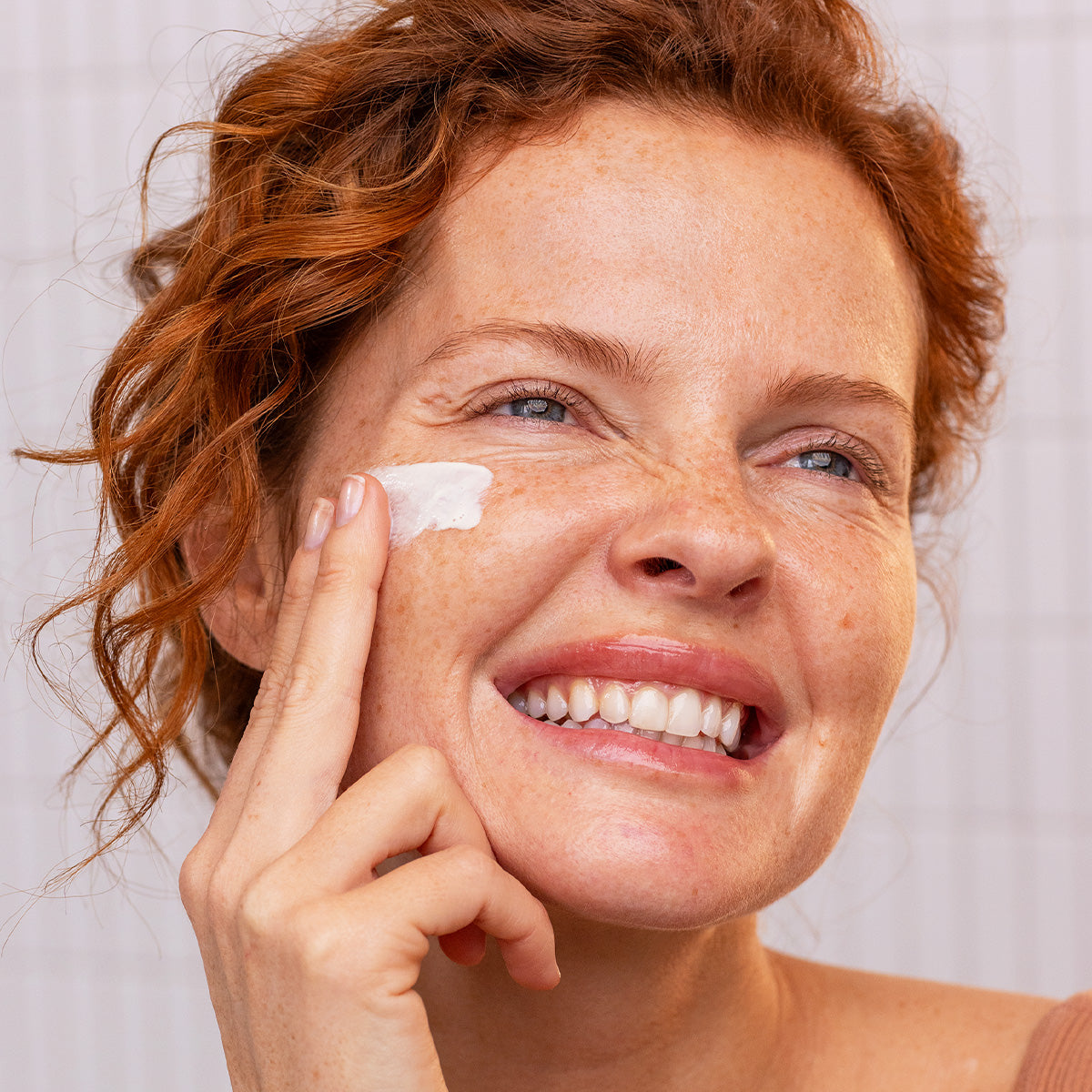 Femme rousse souriante appliquant le fluide solaire Sun Secure SPF50+ sur sa joue, texture légère et invisible.
