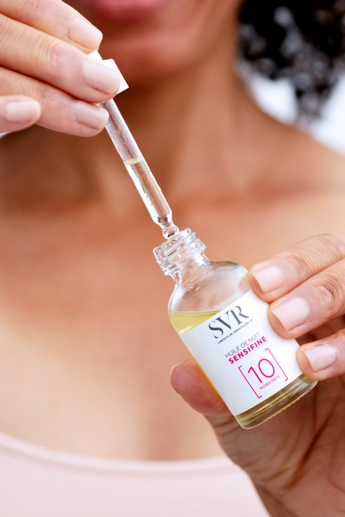 Flacon en verre SVR Sensifine Huile de Nuit avec pipette, tenue par une main, étiquette blanche et texte minimaliste.
