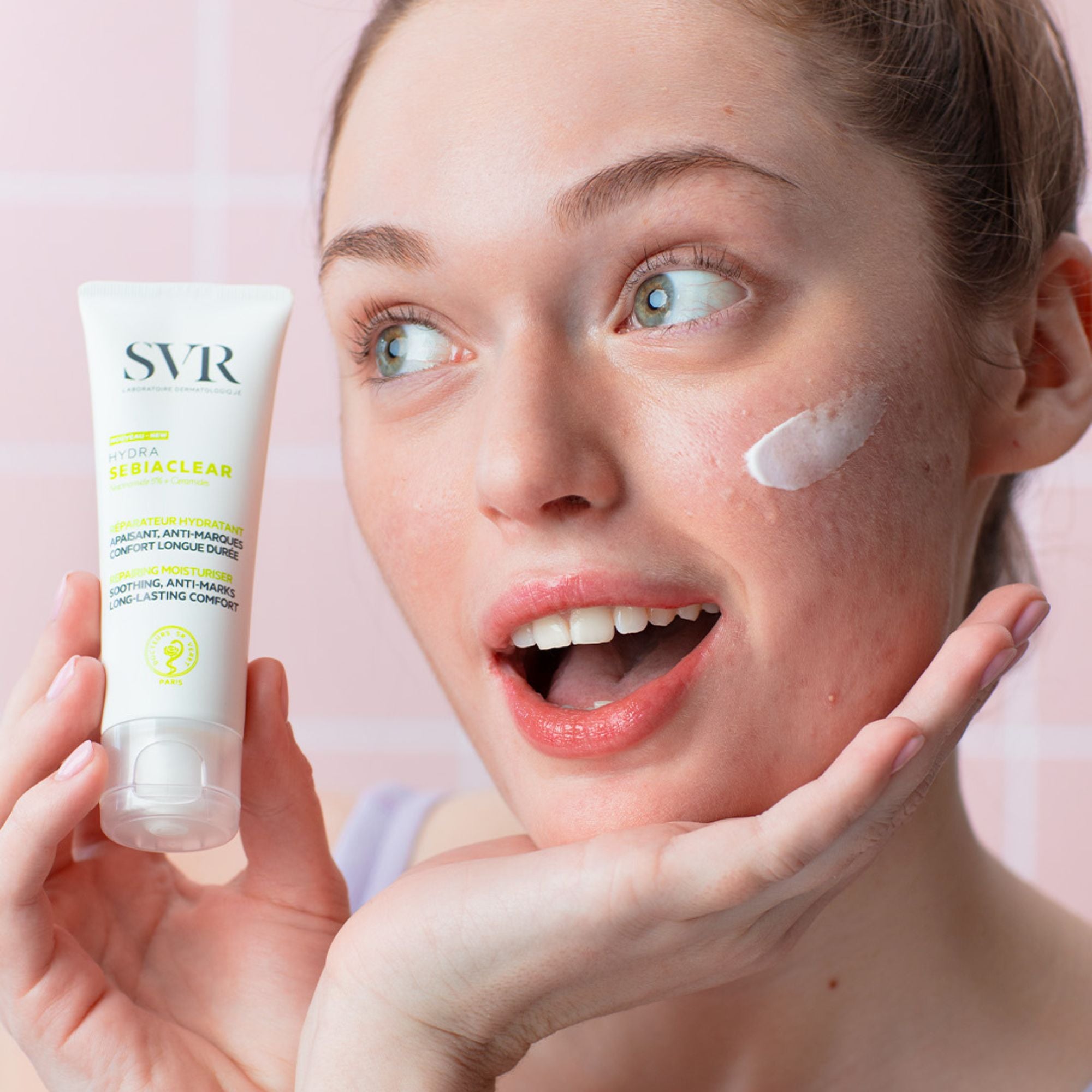 Femme tenant un tube blanc de soin hydratant SVR Sebiaclear Hydra avec une touche de crème sur la joue.
