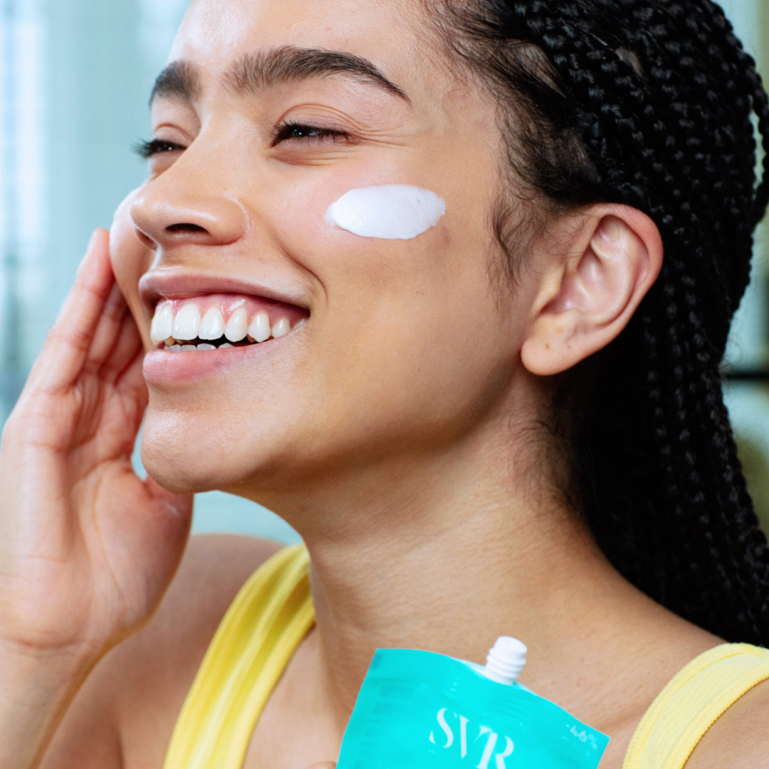 Femme souriante appliquant une crème SVR blanche sur sa joue, portant un débardeur jaune, avec un tube turquoise en premier plan.