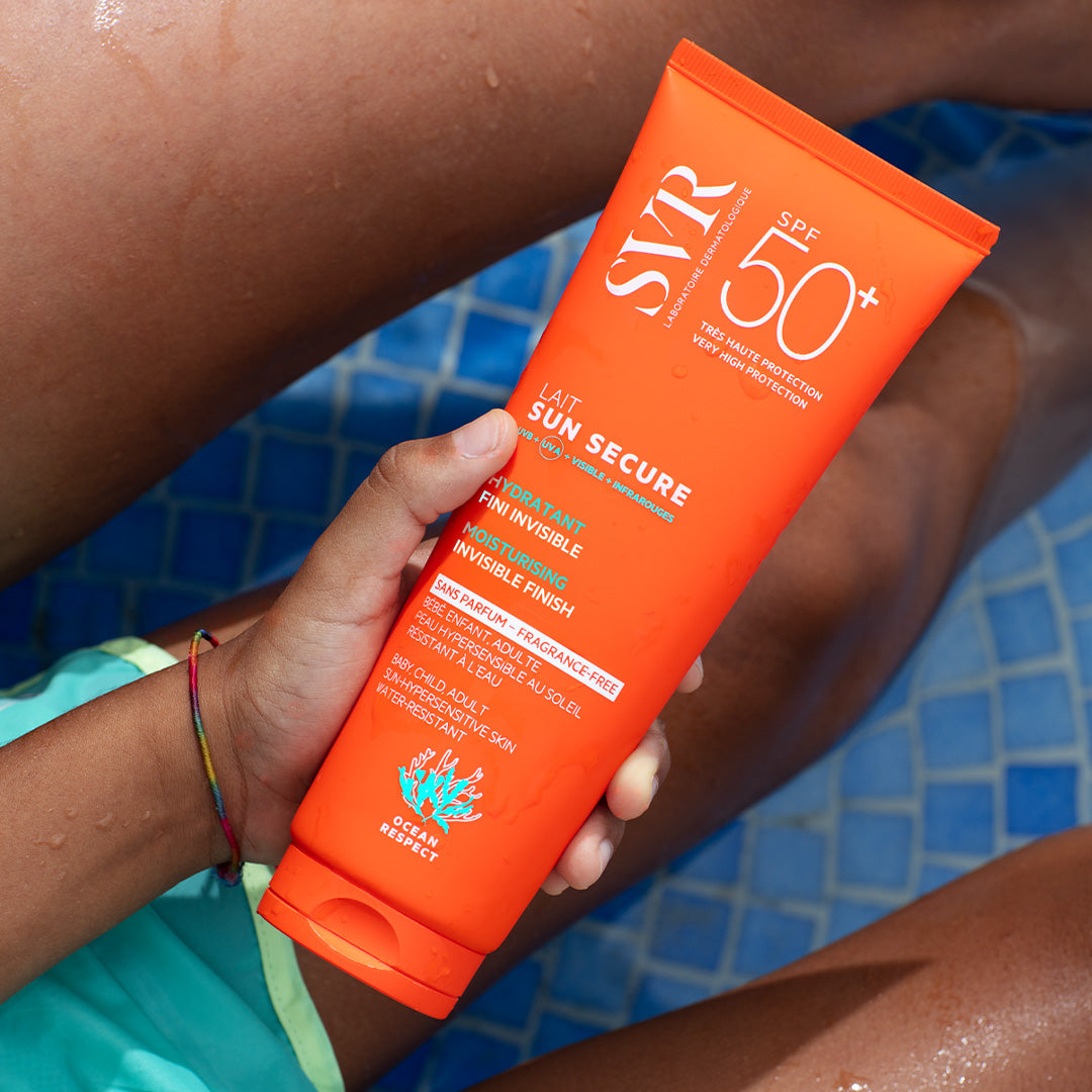 Tube orange vif de lait solaire SVR Sun Secure SPF50+ tenu par une main au-dessus d'une piscine bleue.