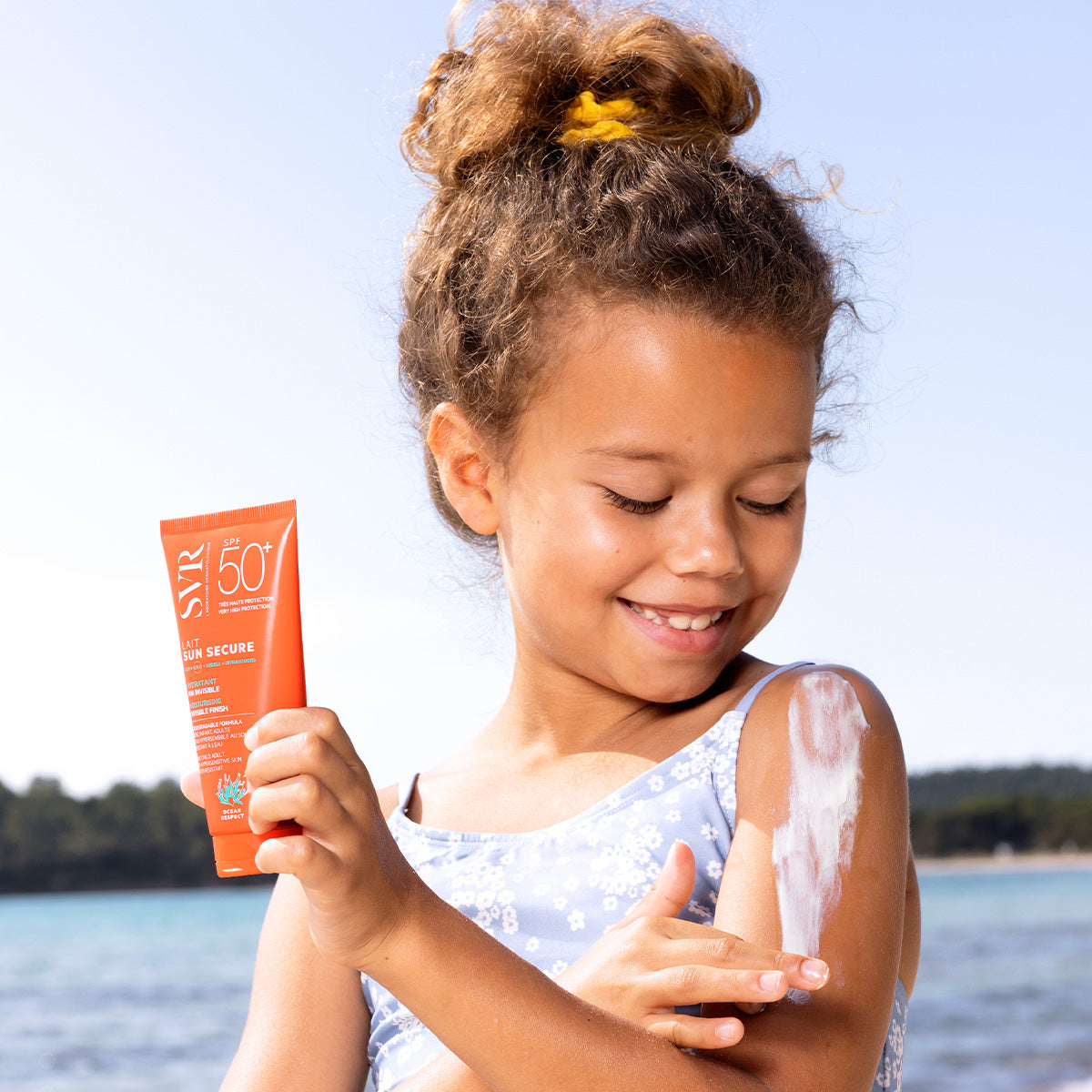 Jeune fille appliquant le lait solaire SVR Sun Secure SPF50+ sur son épaule au bord de la mer.