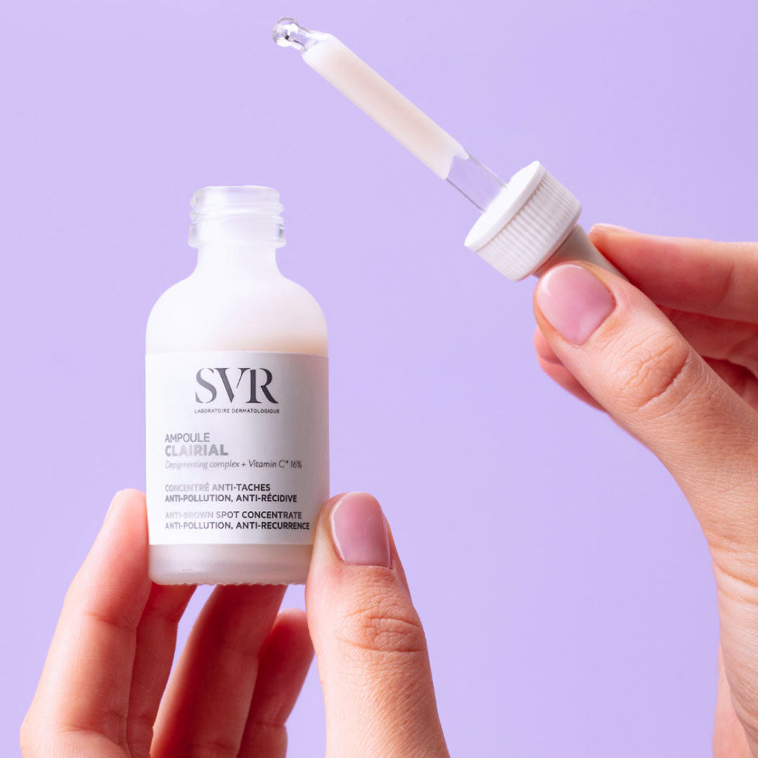 Flacon SVR Clairial Ampoule anti-taches tenu en main avec pipette compte-gouttes sur fond violet.