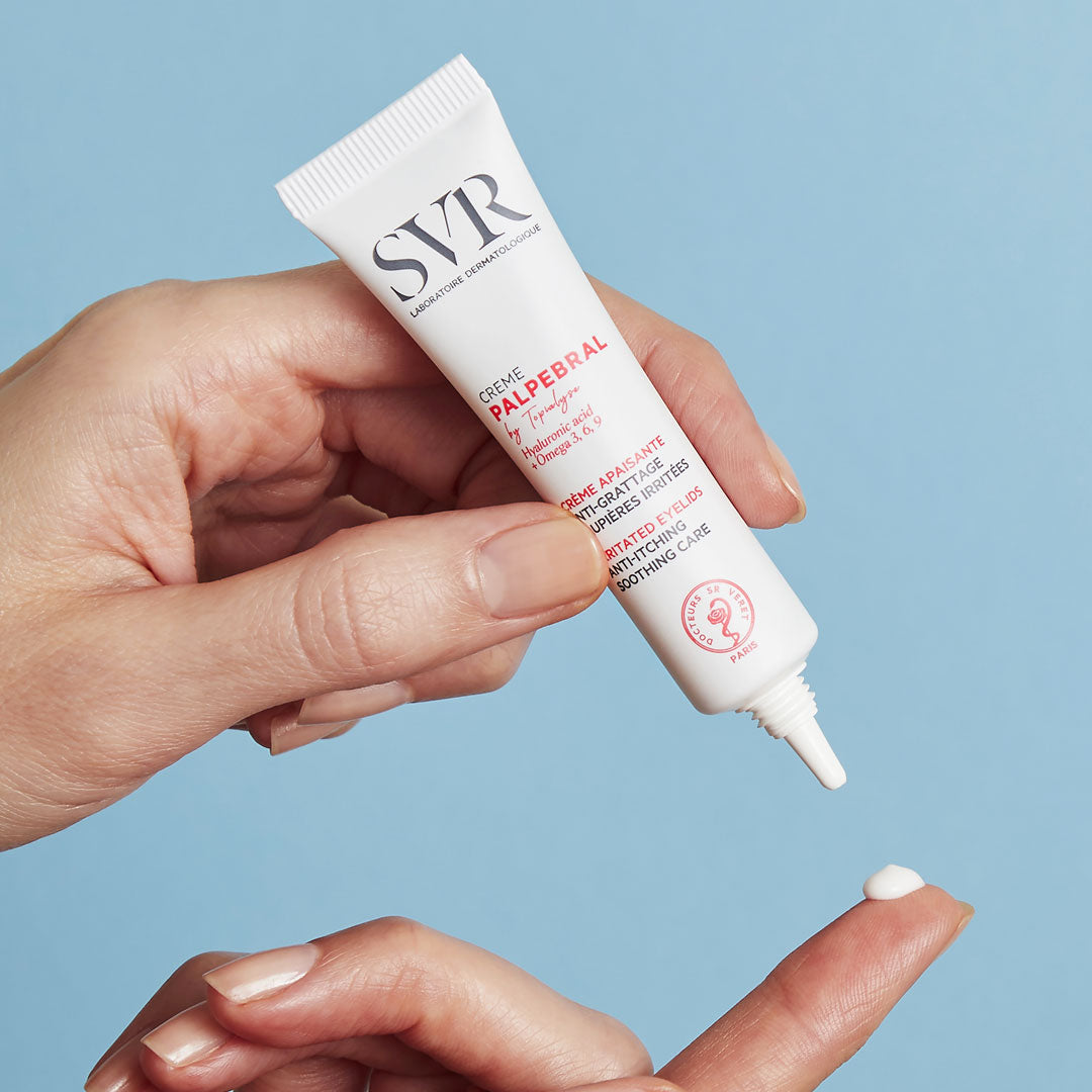 Hand holding a white SVR PALPEBRAL Crème tube, dispensing a small drop of soothing eye cream onto a fingertip.
