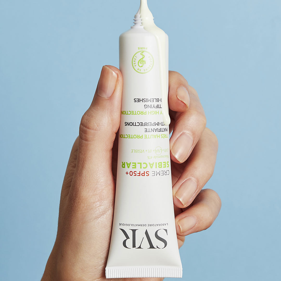 Main tenue en main pressant un tube blanc de crème solaire SVR Sebiaclear SPF50+ sur fond bleu clair.