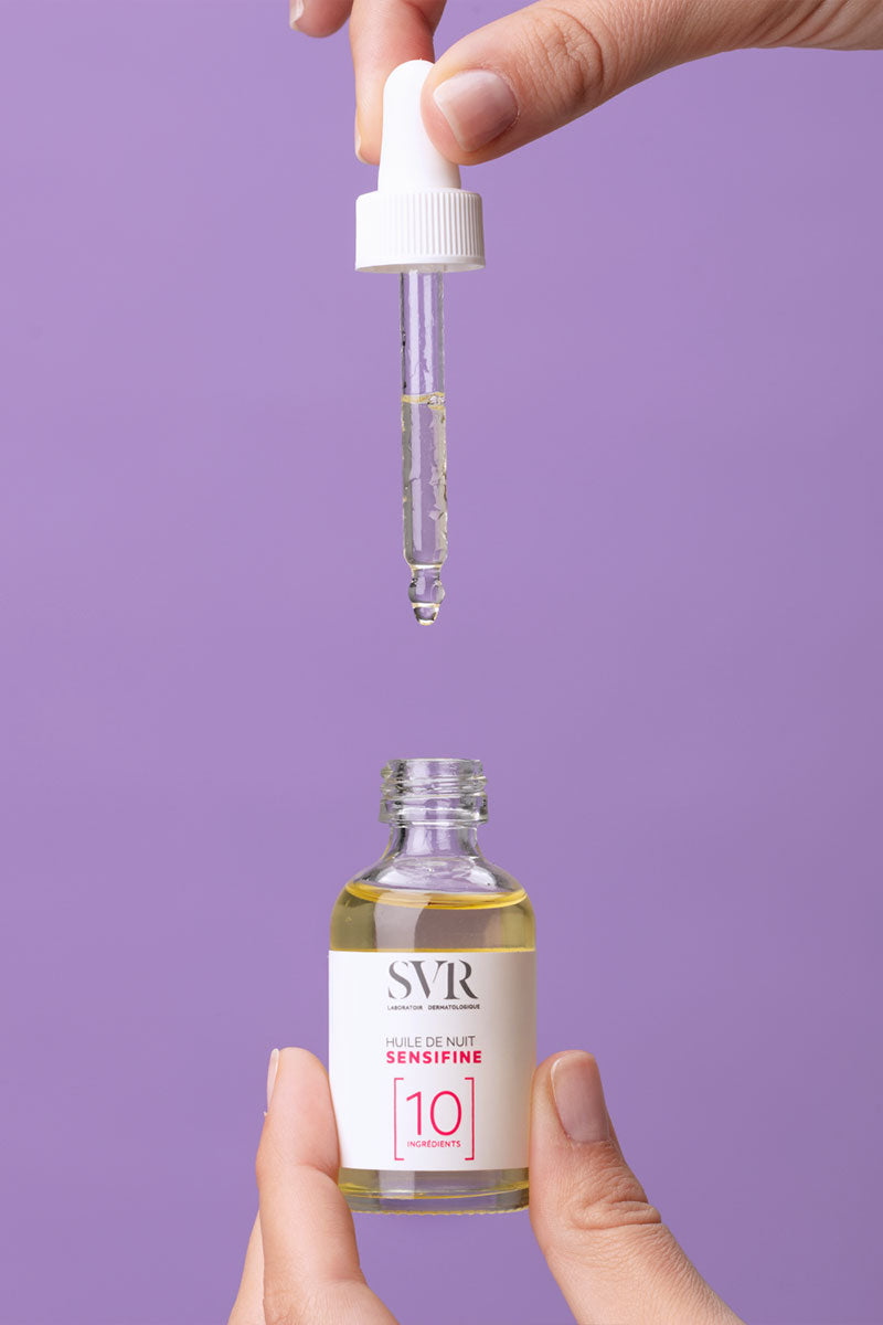 Hand holding a dropper over a bottle of SVR Sensifine Huile de Nuit facial oil against a purple background.