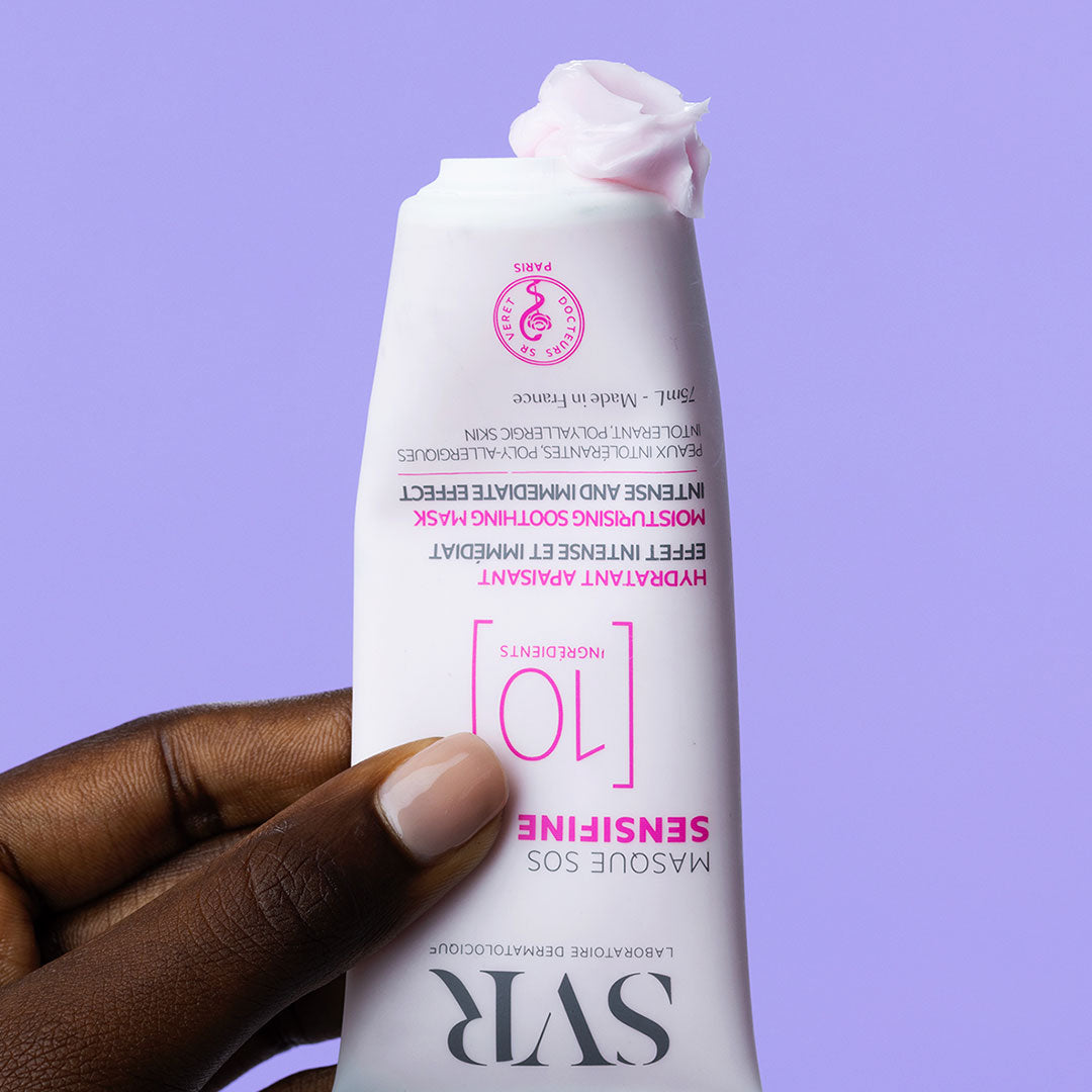 Tube de masque SVR Sensifine SOS blanc avec une noisette de crème rose sur le dessus, tenu en main sur fond violet.
