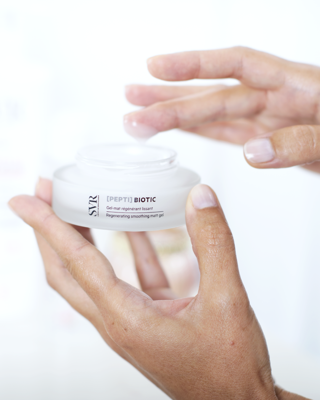 Mains tenant un pot en verre dépoli de gel régénérant SVR [PEPTI]Biotic avec une texture légère sur le doigt.