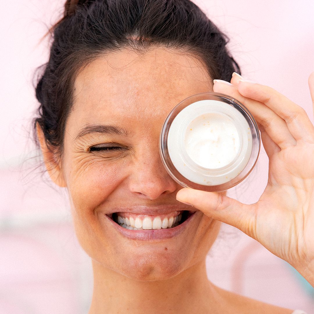 Femme souriante tenant un pot de crème [C20]Biotic devant son œil pour illustrer l'éclat et la régénération de la peau.
