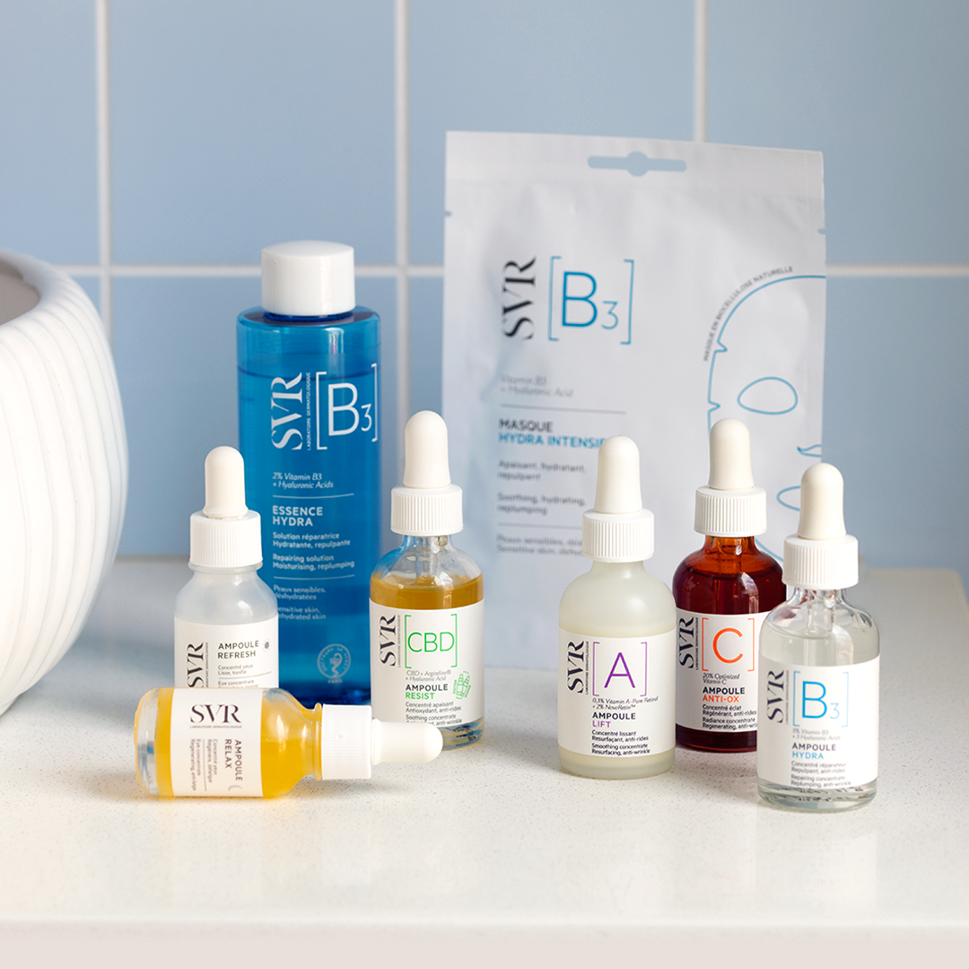 Collection de sérums SVR Ampoule et masque B3 avec flacons compte-gouttes colorés sur un comptoir devant un carrelage bleu.