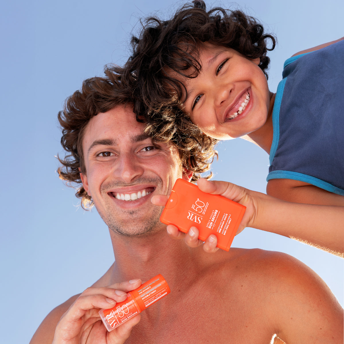 Homme et enfant souriants tenant un spray solaire orange SVR Sun Secure SPF50+ format poche sous un ciel bleu.