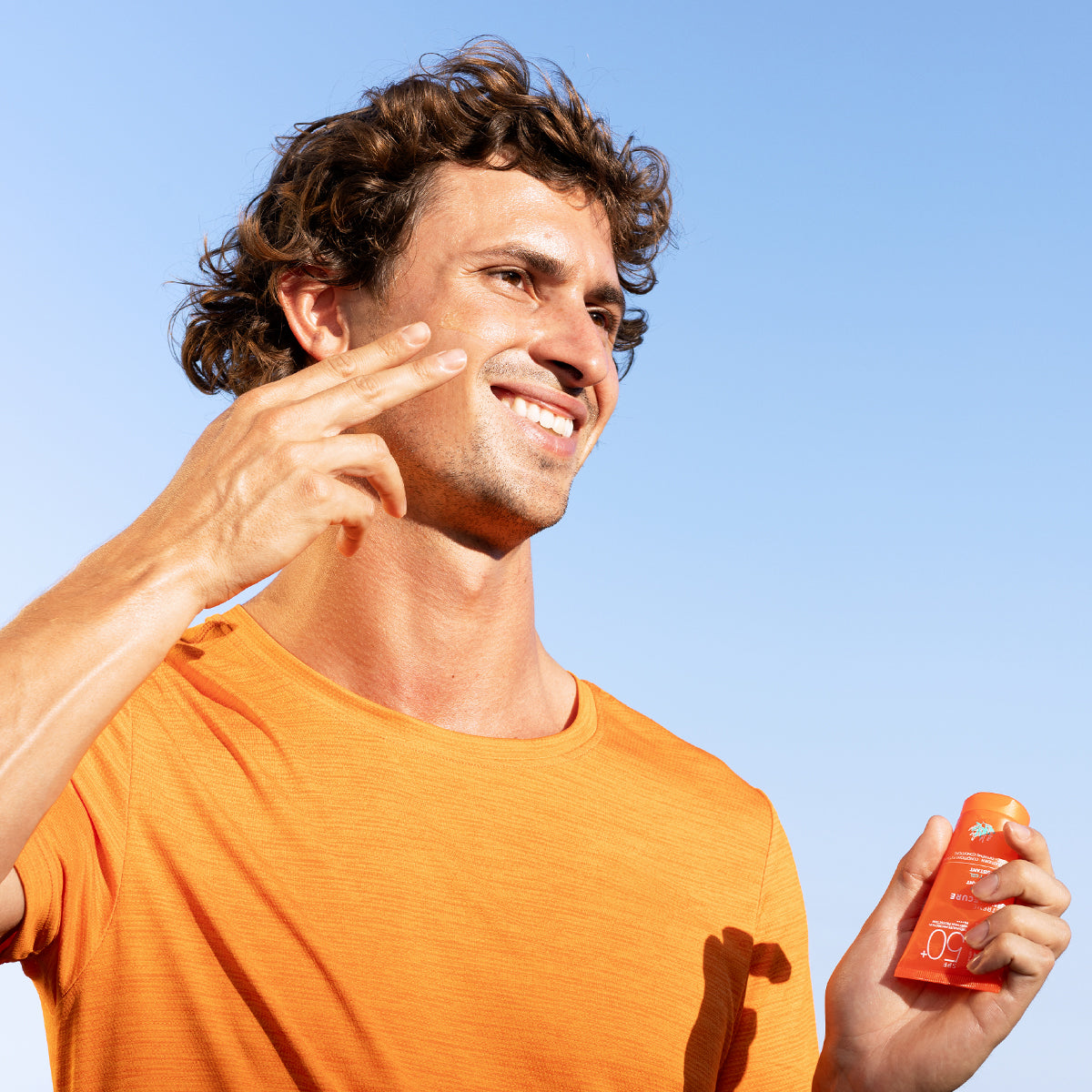 Homme souriant en t-shirt orange appliquant le gel solaire invisible SVR Sun Secure Extrême SPF50+ sur son visage.