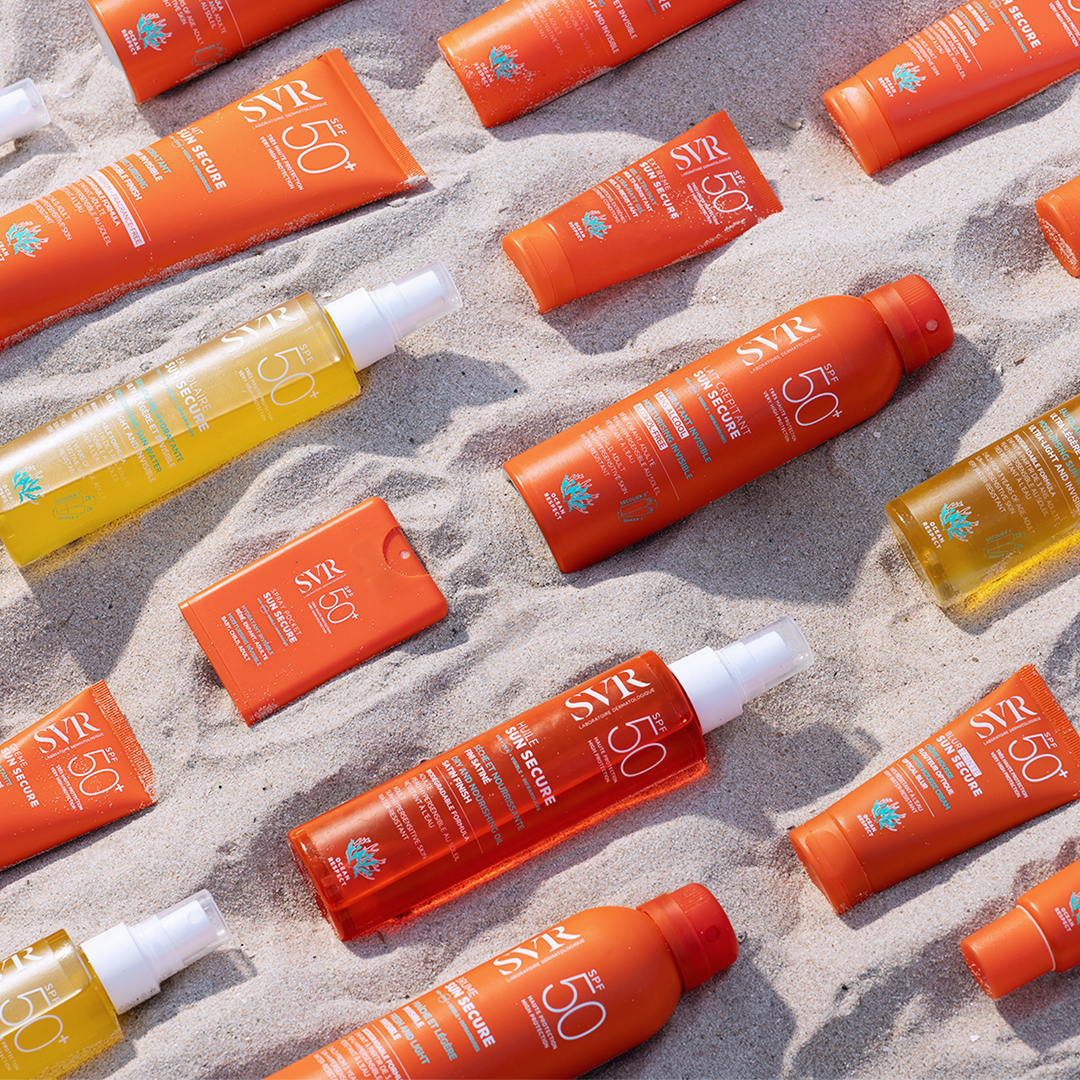 Assortiment de produits solaires SVR Sun Secure SPF50+ aux flacons orange et transparents disposés sur du sable fin.