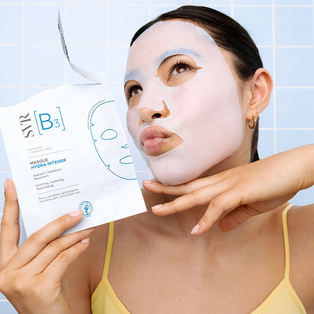 Femme appliquant un masque en biocellulose SVR B3 Hydra Intensif blanc sur un fond de carrelage bleu clair.