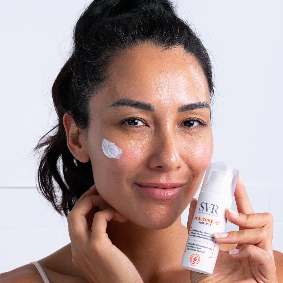 Femme souriante appliquant la crème solaire SVR AK SECURE DM Protect sur sa joue avec le flacon pompe blanc en main.