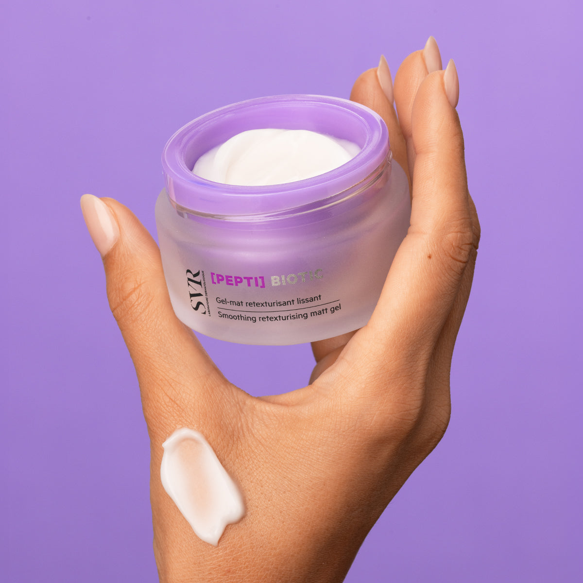 Main tenue en main d'un pot de soin SVR [PEPTI]BIOTIC avec texture gel blanche sur fond violet.