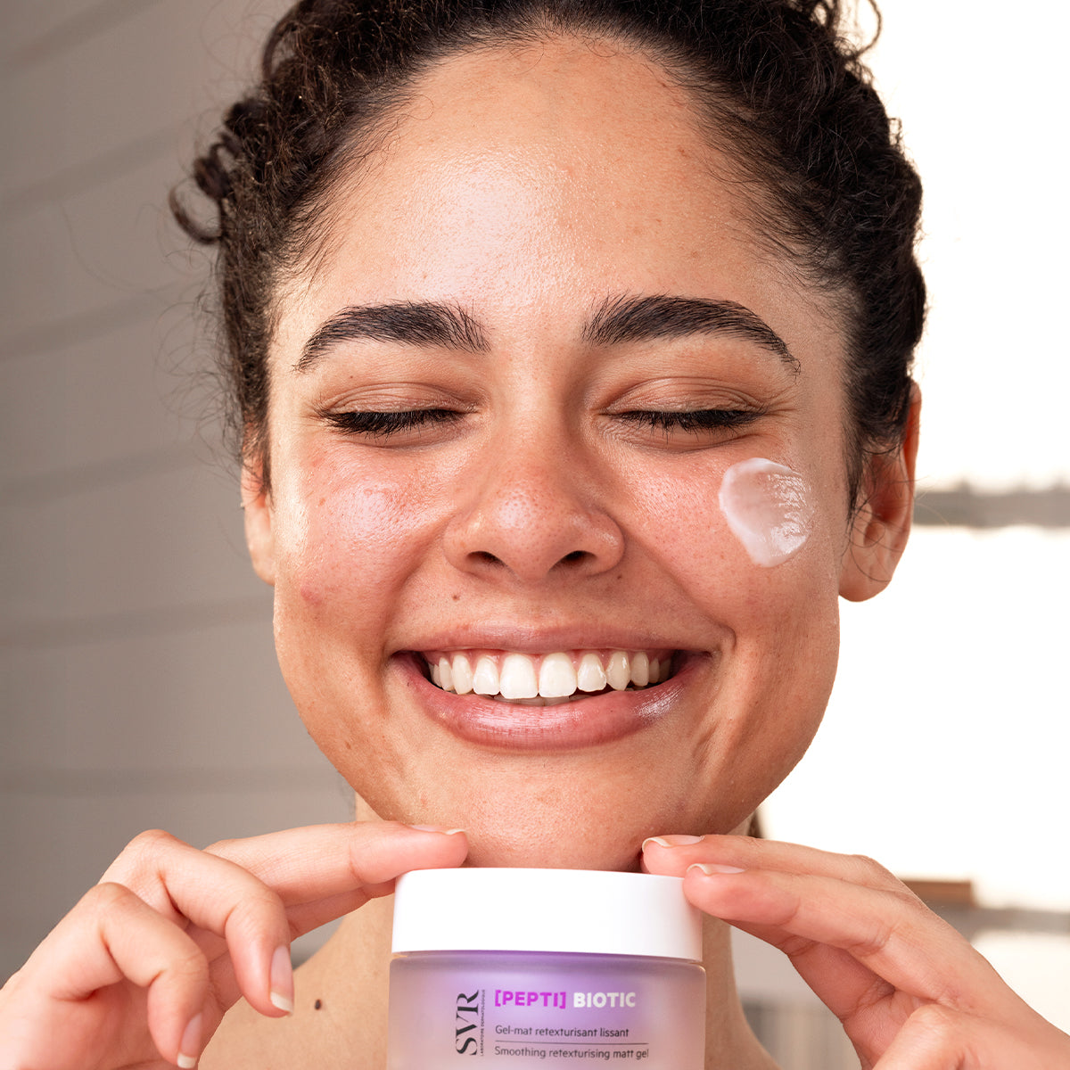 Femme souriante tenant un pot de gel matifiant SVR [PEPTI]BIOTIC avec une touche de crème sur la joue.