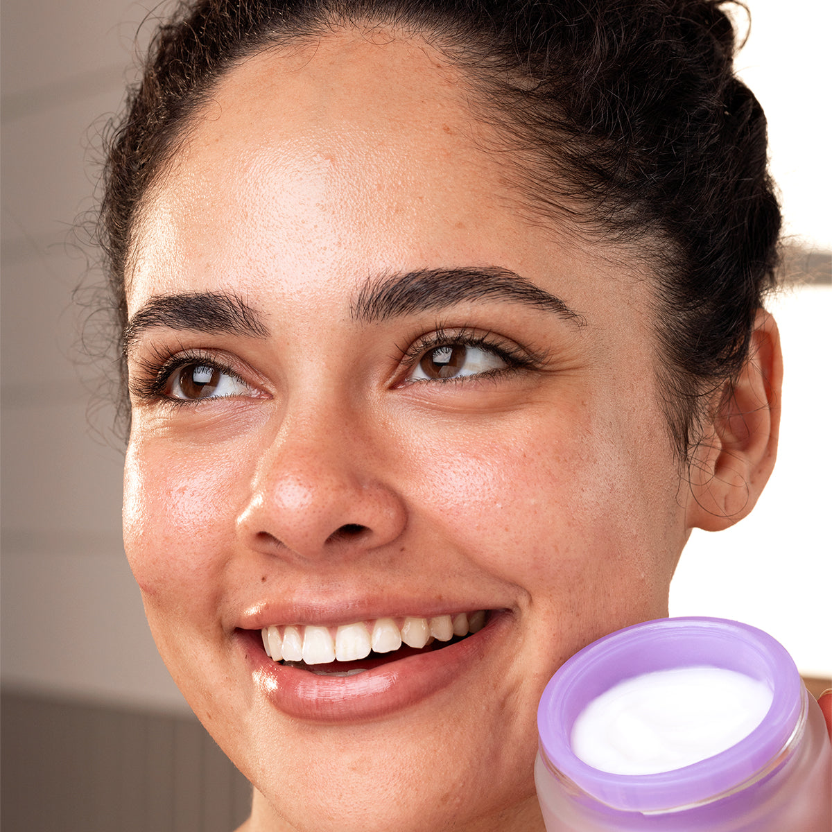 Femme souriante tenant un pot de soin [PEPTI]BIOTIC à la texture gel blanche et au couvercle violet.