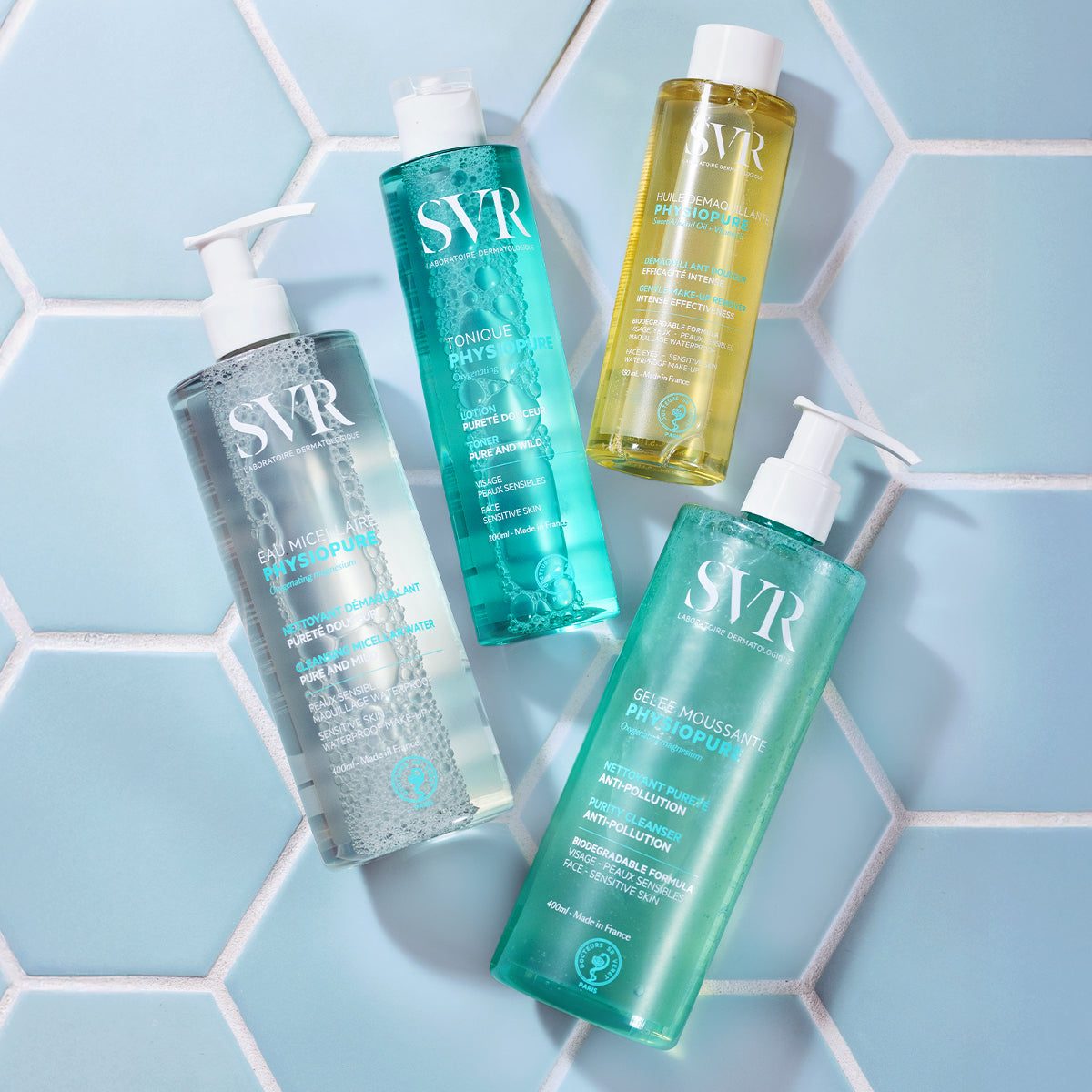 Quatre flacons de soins SVR Physiopure sur un fond de carrelage bleu hexagonal, incluant tonique, gelée et eau micellaire.