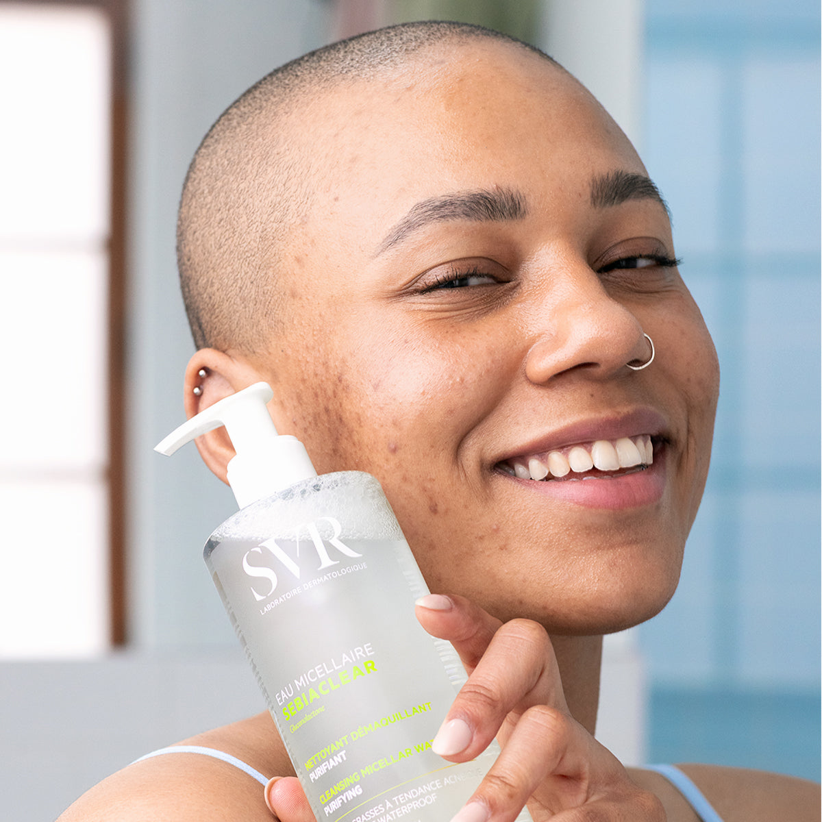 Femme souriante tenant une bouteille à pompe SVR SEBIACLEAR Eau Micellaire transparente pour peaux à tendance acnéique.