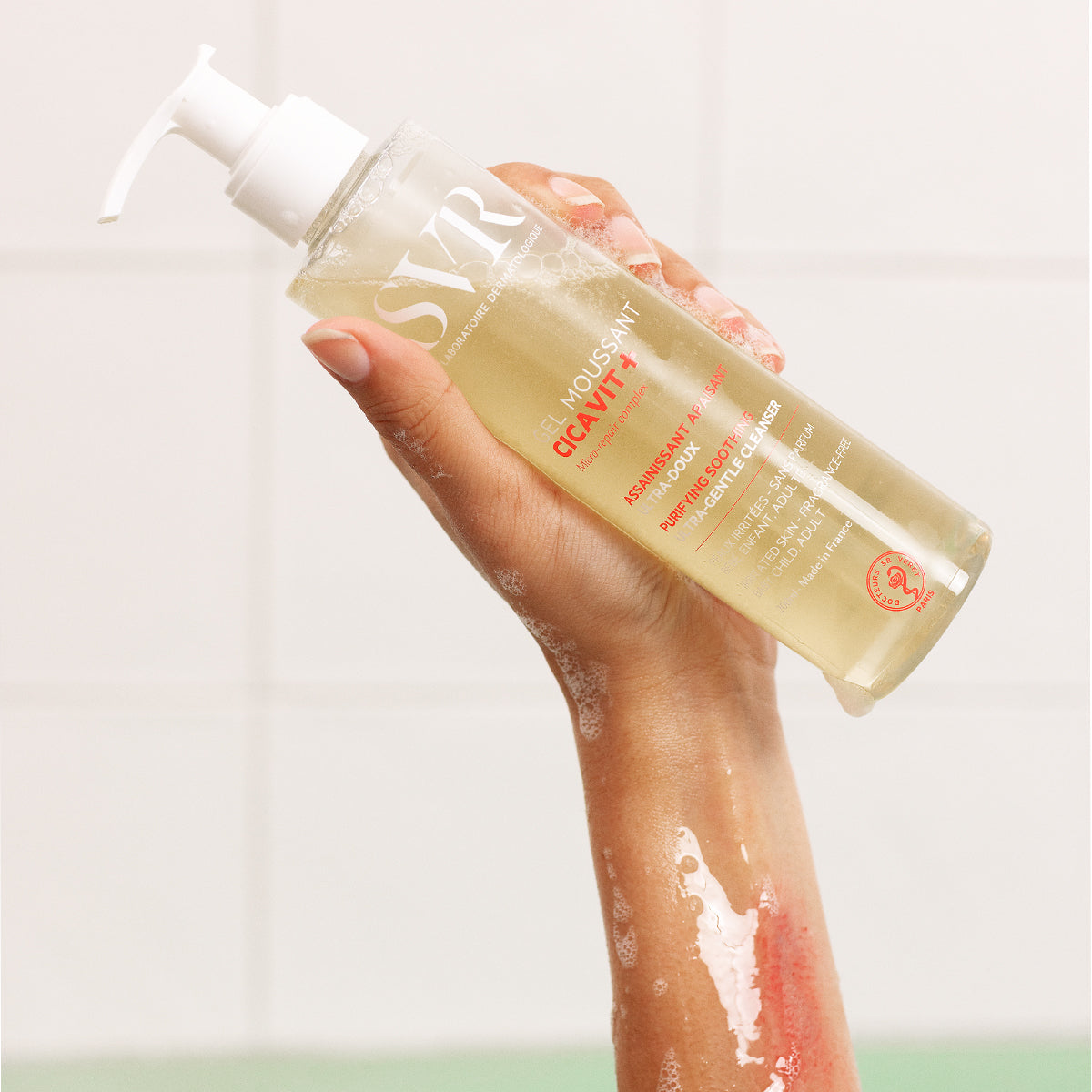 Main tenue par une main, flacon pompe transparent de gel moussant SVR Cicavit+ pour peaux irritées sur fond carrelé.