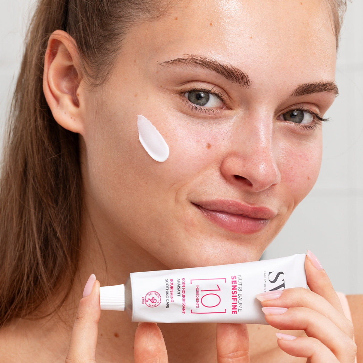 Femme appliquant le baume hydratant SVR Sensifine sur sa joue avec le tube en aluminium en main.