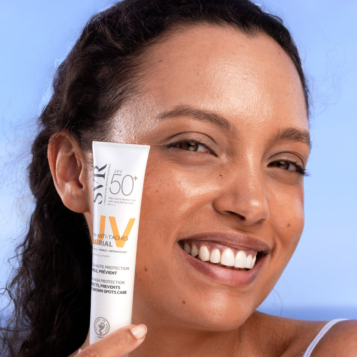 Femme souriante tenant un tube blanc de crème solaire SVR Clairial SPF50+ anti-taches brunes sur fond bleu clair.