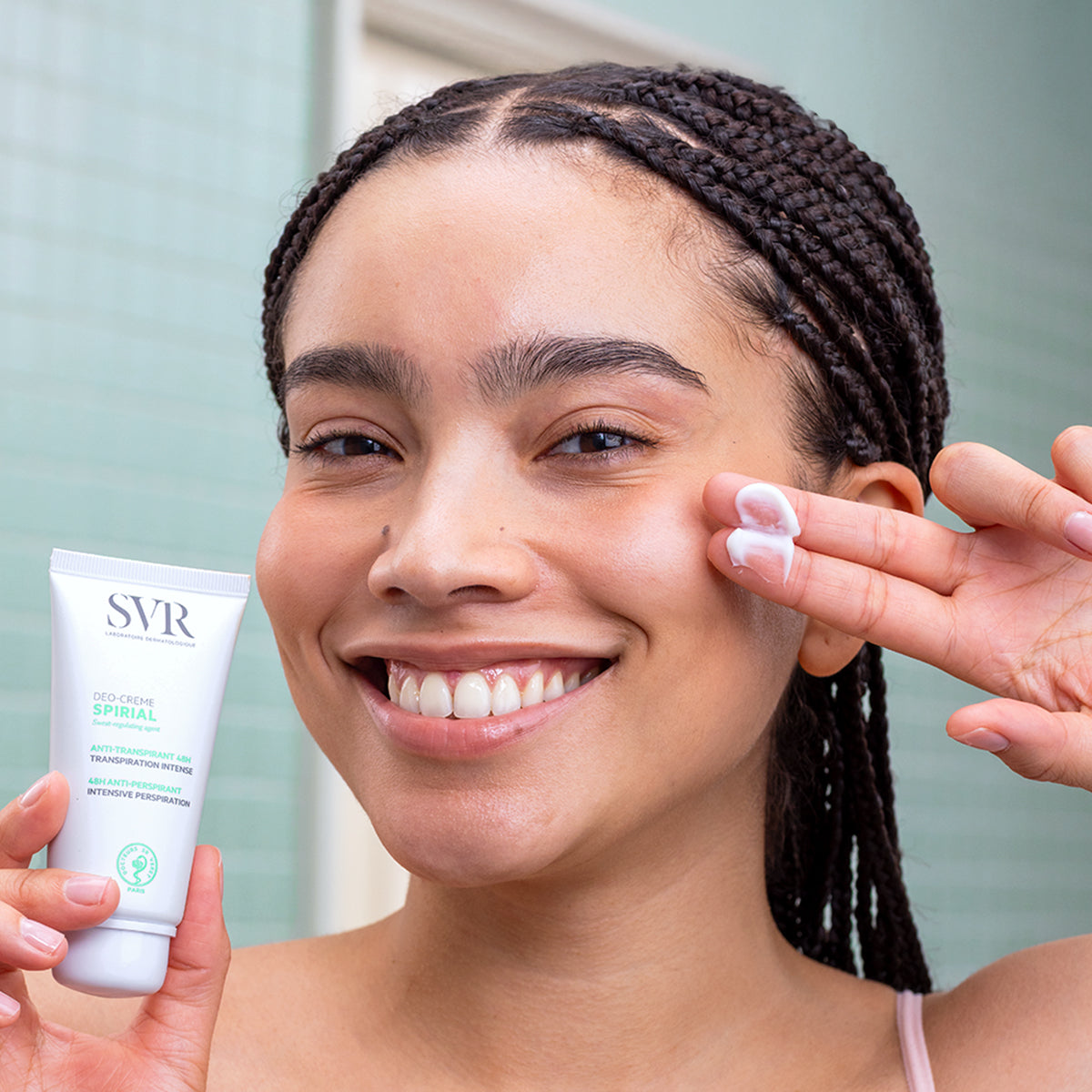 Femme souriante tenant un tube blanc de crème anti-transpirante SVR Spirial et appliquant une noisette de produit sur sa joue.