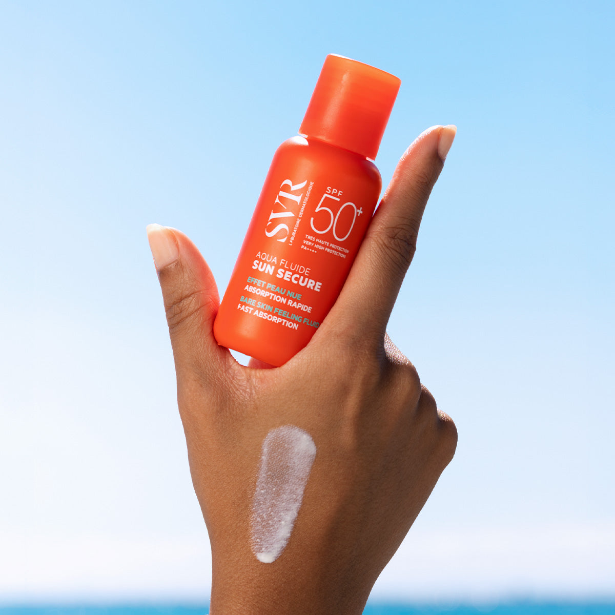Hand holding an orange SVR SUN SECURE SPF50+ sunscreen bottle with a white fluid swatch against a blue sky background.