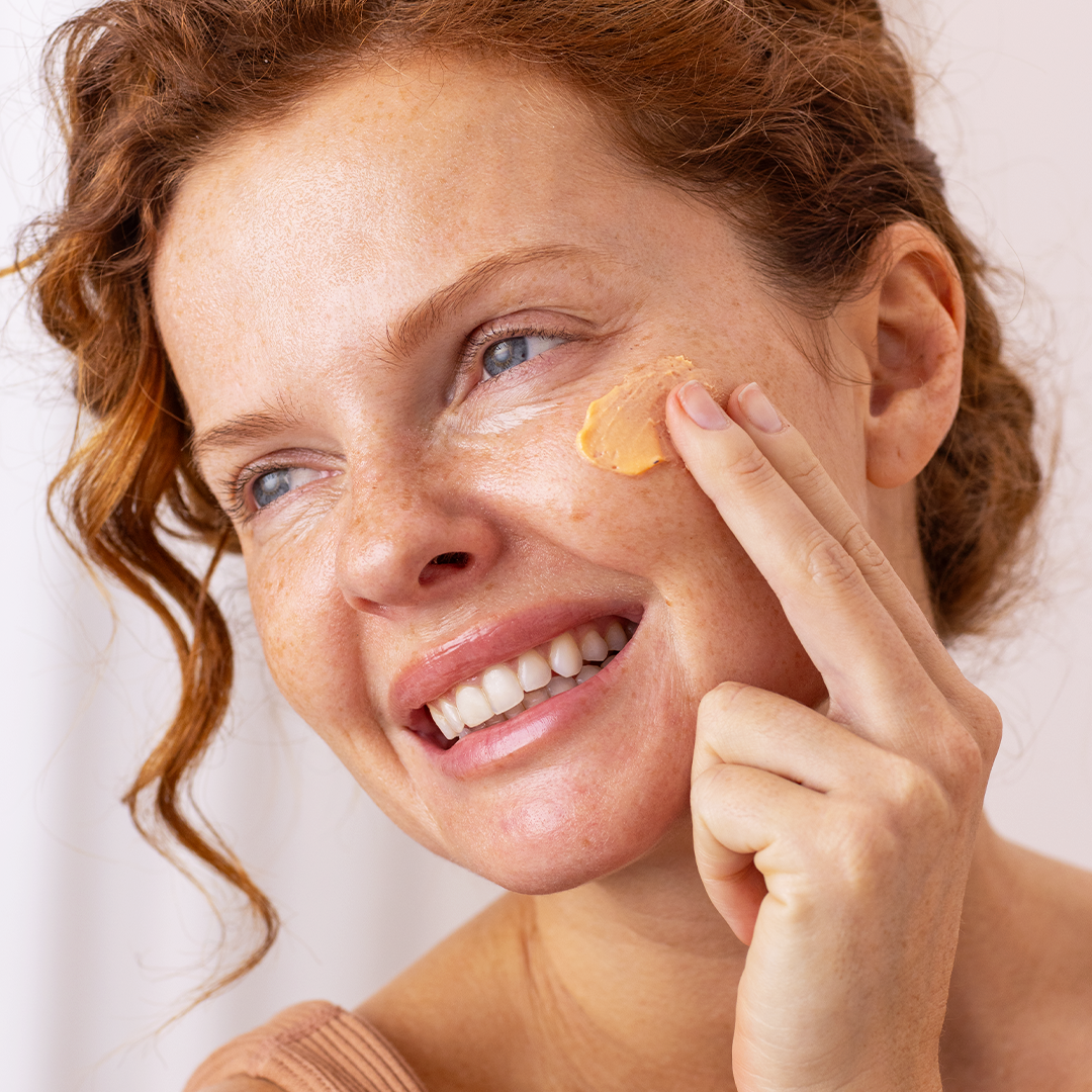 Femme rousse souriante appliquant une crème solaire mousse abricotée sur sa joue pour un teint lissé et velouté.