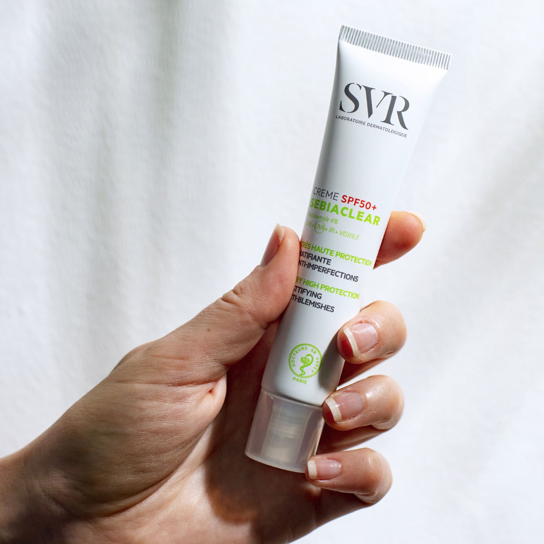 Main tenue en main d'un tube blanc SVR Sebiaclear Crème SPF50+ matifiant anti-imperfections sur fond blanc.