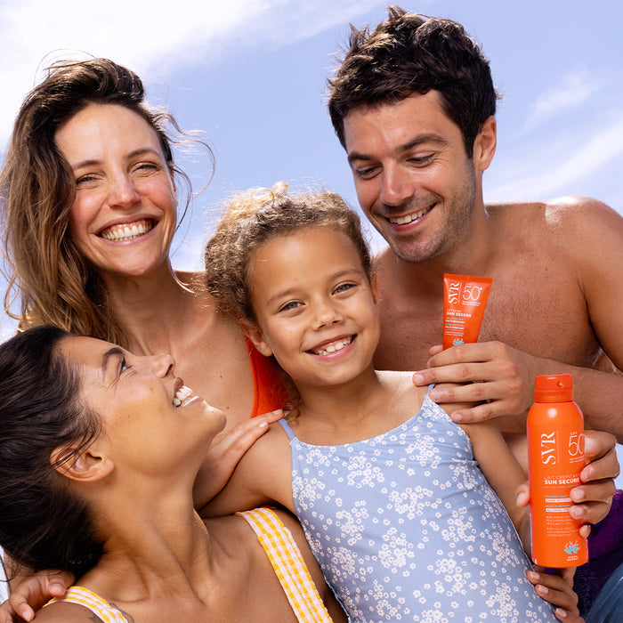 Happy family smiling outdoors while holding SVR Sun Secure SPF 50+ sunscreen products against a bright blue sky.