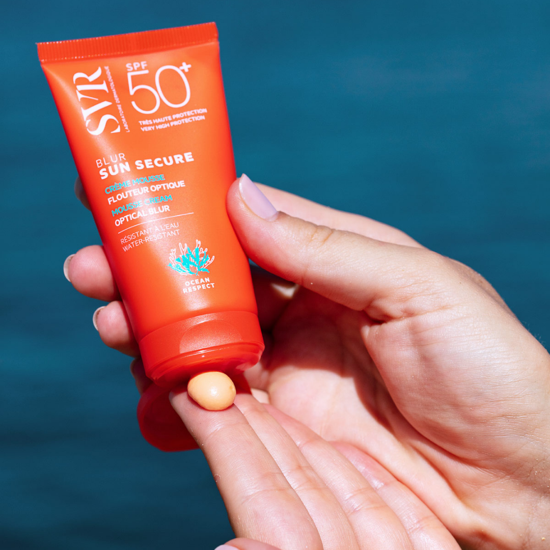 Tube orange de crème solaire SVR Sun Secure SPF50+ pressé sur un doigt, révélant une texture mousse abricotée sur fond bleu.