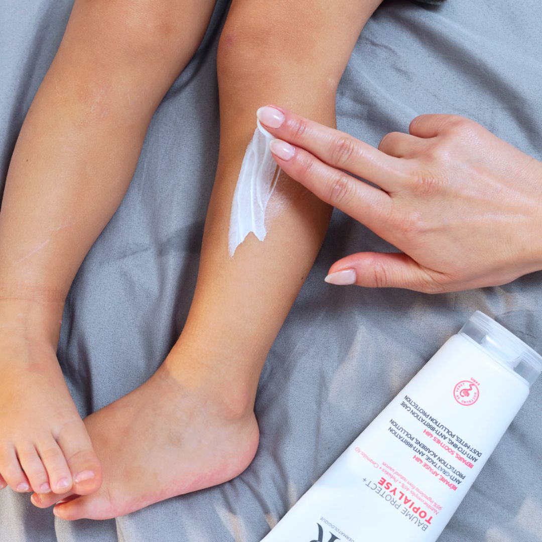 Main appliquant le baume réparateur TOPIALYSE Protect+ sur la jambe d'un enfant posée sur des draps gris.