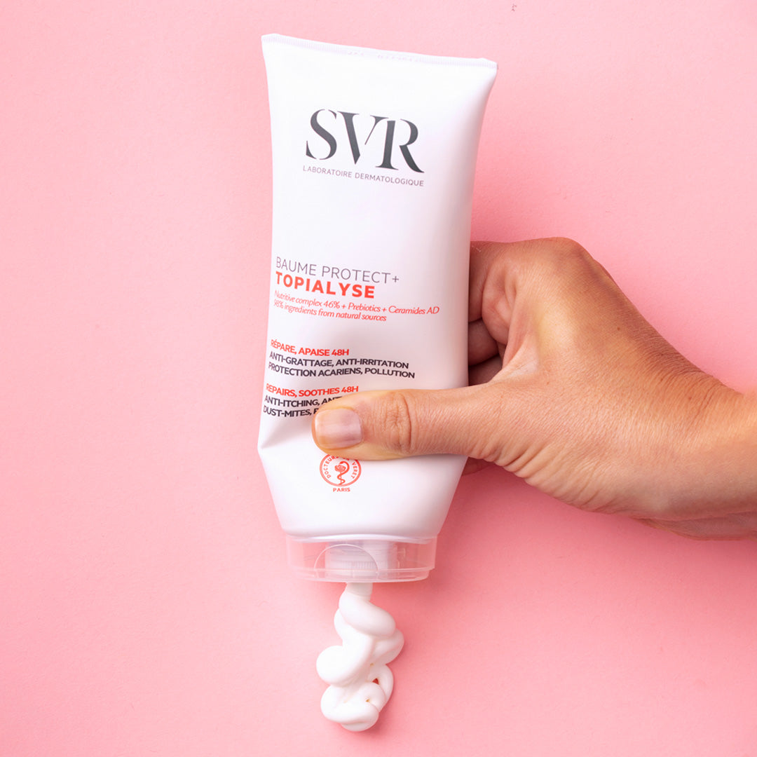 Main SVR TOPIALYSE Baume Protect+ pressé à la main, délivrant une texture crème blanche sur fond rose.