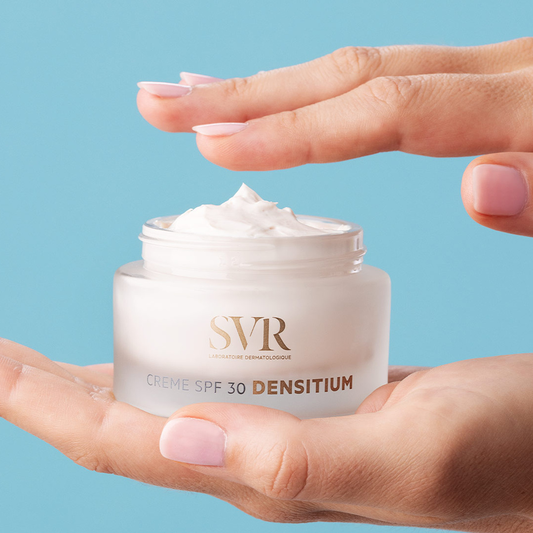 Hands holding an open jar of SVR Densitium SPF 30 cream with a smooth white texture against a light blue background.