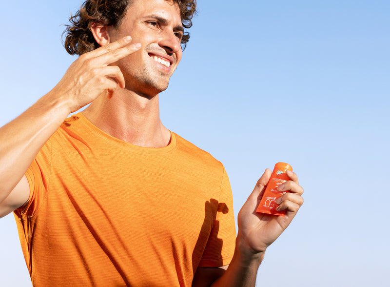 Smiling man in an orange shirt applying SPF 50+ sunscreen to his face against a clear blue sky.